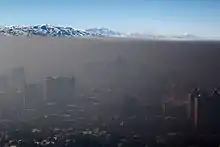 Dense, smokey air lays over a cityscape like a blanket. In the distance, a mountain range and clear blue sky can be seen.