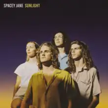 The four band members standing in front of a purple and yellow gradient, looking up to their right.