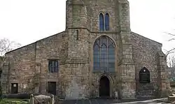 Stone building with two sloping wings, the left wider than the right; large stained glass window above door in the center, topped by lower part of tower