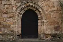 A small window with four leaded panes in a rectangle, another thin pane to the left of thee, in a worn stone-like frame, surrounded by sand coloured stones