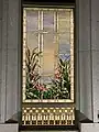 A stained glass window from the Orem Utah Temple, depicting bullrushes and cherry blossoms, next to water elements, with a blue sky with clouds. It is framed by a golden rectangular frame, and has stone masonry surrounding the window.