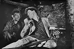 Alexei Stakhanov and another man at work in a Soviet coal mine. Stakhanov, while holding a drill, is seated at the coal face, his head turned to speak to his colleague.