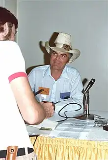 Man in a cowboy hat and white shirt with thin blue stripes sitting at a table with a microphone