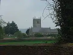 Gray stone building with square tower behind. In the foreground are green fields and bushes.