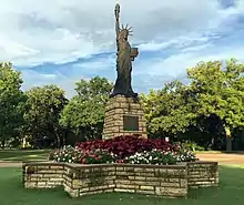 Photo of a Statue of Liberty scale model replica, about 20 feet tall. It is situated on its pedestal which stands within a star-shaped basin made of limestone pavers, which in turn acts as a planter full of a lush arrangement of multicolored flowers and foliage. The sunlight dawns upon the statue's face, with a background of a treeline, and a cloudy blue sky.