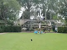 a grassy lawn in front of a statues complex several people sunbathe or sit in the midground facing in the background a group of white statues that are significantly larger than the people. In the foreground a black bird is walking away from the whole tableau