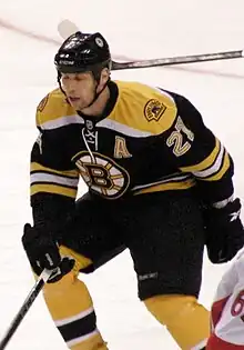 A hockey player in a Black uniform with gold trim and a stylized "B" logo on his chest defends his position during a game.