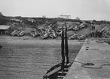 View of a pier leading to a sloping shingle beach on which numerous boats are standing. Groups of people are visible at the foot of the pier and on a track which leads up to a building at the top of the slope.