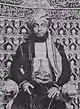 A black-and-white photograph of a man with a dark beard wearing a turban, a dark jacket, and a white shirt, sitting, and looking at the viewer