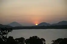 A partial view of a large lake, seen over a tree line. The further bank is covered by a thick forest, and the sun is setting over a mountain range in the background.