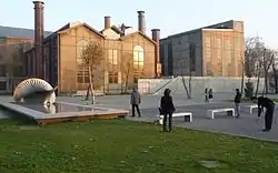 A brick factory stands in front of a park, with open green space, a reflecting pool, and benches