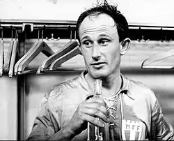 A dark-haired gentleman in light-coloured football kit stands in a dressing room, holding a bottle in his right hand.