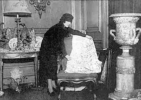 White woman in smart day clothes, including a hat, in a shop full of antiques and objets d'art