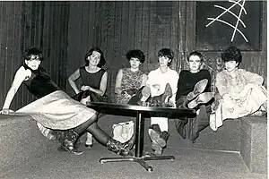 Six dark haired women sitting at a table in a black room