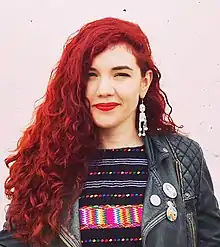 A women with curly red hair stands in front of a pink brick wall.