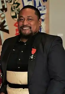 a man with short black hair, a tidy beard and moustache smiles directly at the camera. he has a black jacket with a medal and a black shirt. He is also wearing a fine mat woven from natural fibres