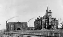 Two buildings stand side by side on a hill. The one on the left is two stories, with two smokestacks behind it. The one on the right is the larger, taller Tech Tower building.