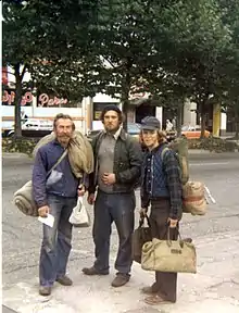 Ted Conover in early 20s with sparse beard, collar-length hair, cap, and hobo traveling clothes and gear, standing on a sidewalk with two other railroad hobo men carrying gear