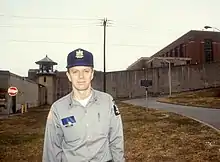 Ted Conover in his late 30s, standing outside of the prison perimeter wall wearing his correctional officer uniform hat and button-up shirt, with officer's notebook and pen in his shirt pockets