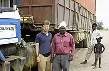 Ted Conover stands with a Kenyan man next to a large industrial truck parked on a dirt road while a younger Kenyan man and little boy look on.