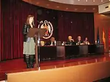 Woman speaking onstage, with others seated at a dais