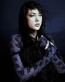 Teya Dora wearing a dark blue patterned top, arms crossed in front of her chest, looking forlorn to the left of camera