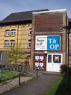 The image shows the ThOP's audience entrance. It is a tall cuboid containing a staircase. Behind that, a flat cuboid with a regular European rooftop, houses the actual theater.