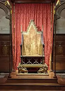 An old gilded wooden chair on a raised platform in front of a red curtain.