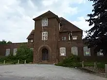 A photograph of a large derelict brick building from the 1930s. The front of the building has a large, rounded archway in the art deco style. The lower windows are boarded up, and the panes of many of the upper windows are smashed