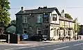 The Plough, opposite the station site, which acted as ticket office before the station was built. The footpath to the left, between two fences, follows the course of the dismantled line.