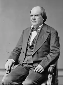 A man with dark hair, bald on top, wearing a black suit and tie with a white shirt, seated