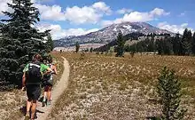 Two hikers with backpacks walk along a trail in the Three Sisters Wilderness, surrounded by trees with the Three Sisters visible in the background.