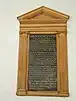 A photograph of a memorial in a wooden frame hanging on the wall of a church. The frame is a carved depiction of a Greek temple with columns and a small gable. The inscription is of gold lettering on a black background