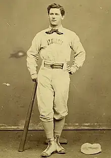 A baseball player is standing, facing the camera, with the top of a baseball bat in his right hand, and the bottom resting on the ground