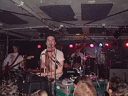 A band play in a crowded room. A man is playing keyboards while singing into a microphone; behind him is a drummer and to one side is a guitar player.