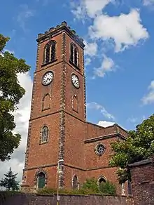 A tall slim tower with clock faces and a battlemented parapet seen from below its foundation on a nearby street