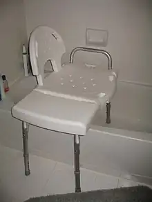 A white plastic bench sits with two legs inside a bathtub and two legs outside the tub resting on the floor.