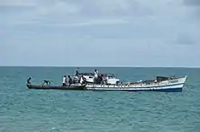 Seven or eight people work to load logs from a smaller, wooden vessel (carved from a tree trunk) onto a slightly larger boat