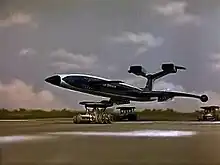 A blue, futuristic airliner rests on top of three orange support vehicles (one underneath the fuselage and two underneath the wings) as it speeds out of control down an airport runway, the sky bright with clouds.