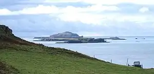 Brown-coloured islands of varying elevations lie in a calm sea beneath cloudy skies beyond a grassy slope.