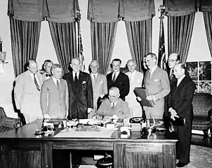  Eleven men in suits stand around a large desk at which another man is signing a document.