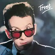 A headshot of a man wearing a suit and red-tinted glasses staring up with his eyebrows perked up. The word Trust appeared underlined in the upper right corner.