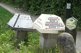 Four bike signs near the Dutch-German border; a Dutch mushroom with a metal cap, a German mushroom with metal plates, a flat named-route sign, and an internode sign.