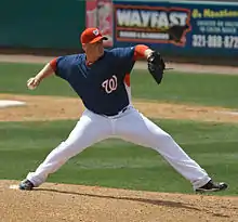 A man wearing a red baseball cap, navy-blue baseball jersey with a curly white "W" on the left breast, a red undershirt, and white baseball pants throwing a baseball with his right hand