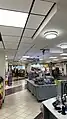 A patterned kitchen-island with a container of water bottles, two short racks of chip bags, and a double sided soda dispenser sitting on top. In the rear are students waiting in front of food booths. A small sign on the ceiling reads "Coffman Market Beverages and Cashier.