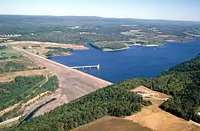 An aerial view of a large dam and lake surrounded by forest with low mountains in the background