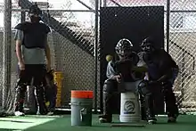 A man with a full set of catcher's gear sits on a bucket with his glove out as a baseball heads towards him in a bullpen. Behind him is an umpire and another catcher observing from the side.