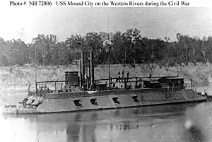 Black and white photo shows a warship with a sloped casemate and two smokestacks. There are four broadside guns and three guns forward.