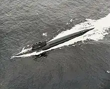 A surfaced submarine is seen from above and to port making high speed, with a long wake around and behind.