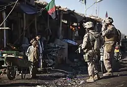 Two Marines stand off to the side of a road near several dilapidated buildings while talking to a group of small children.